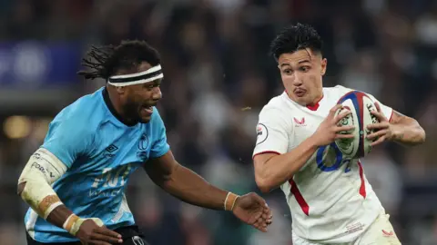 Elia Canakaivata (left) playing for Fiji, wearing a white headband, attempts to tackle England's Marcus Smith who is carrying the rugby ball