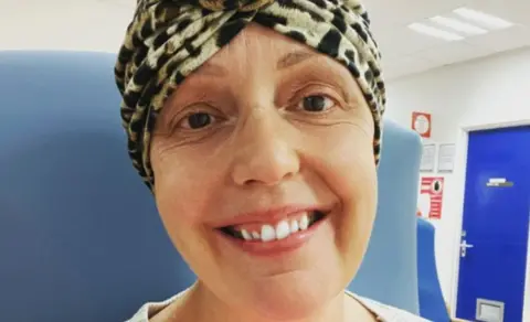 Hollie McFarlane Hollie sat in a hospital chair wearing a head scarf in hospital. She is smiling at the camera