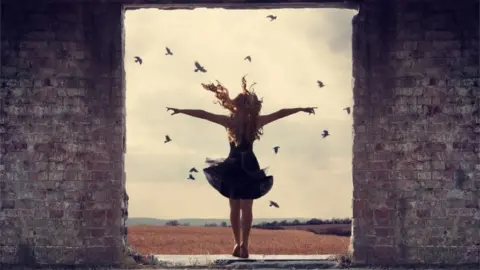 Joanna Wilson A woman looking out at a view with her arms in the air with birds in the sky