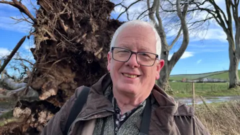 Bob McCallion is smiling into the camera. A large tree is lying behind him.