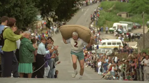 Getty Images A man with grey hair and a bib with the number two is running up a hill with a large brown sack on his shoulders. The street is lined with crowds of people.