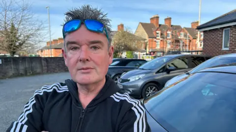 BBC Gary Davies berdiri di tempat parkir. Ada mobil abu -abu di belakangnya. Dia mengenakan kacamata hitam dan atasan pakaian olahraga Adidas. Dia memiliki rambut spikey. 