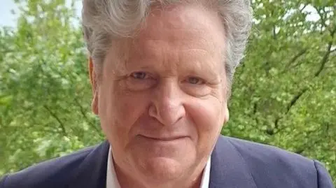 Rejoin EU Party John Stevens smiles for the camera. He has thick grey hair and is wearing a blue jacket and open necked white shirt.