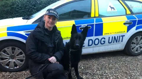 A photograph of a female police officer kneeling next to a black German Shepherd dog. There is a "Dog Unit" police car in the background.