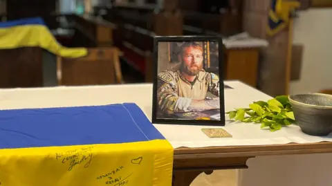 BBC A framed phot of Chris Garrett in military fatigues placed on the alter with the blue and yellow of the Ukrainian flag in the background.