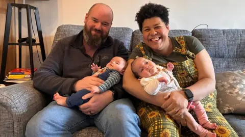 Elita and Adam sit on a couch, each holding a baby. Adam wears a dark jumper, and Elita wears a bright sunflower-patterned overalls. A lamp and stacked books rest on a small table beside them, creating a warm, homey scene.