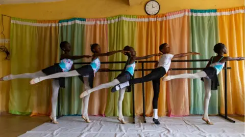 Young dancers dressed in matching leotards hold a ballet barre during a class.