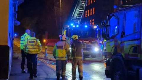 Firefighters and other responders in high-vis jackets stand in front of a burning building. There is a fire engine with its ladder raised and another engine closer to camera. it is night.