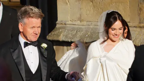 PA Media Gordon Ramsay pictured entering Bath Abbey with his daughter Holly. He is wearing a black suit and she is wearing a white dress and veil. 