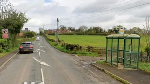 Google A rural road which has a green field on one side of a road. Next to the field is a bus shelter. There are two cars on the road and arrows and road markings. 