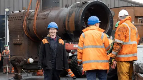 KWVR A woman in blue hard hat and black and orange donkey jacket standing in front of a rusting locomotive's boiler being lifted by a crane