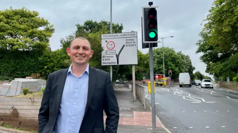 A man in a dark blazer and blue shirt is smiling, and stands in front of a road sign detailing a junction. The road on which the sign stands is lined by trees.