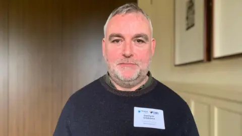 BBC A bearded man with grey hair in a blue jumper with a green shirt showing underneath