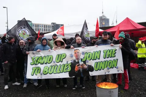 PA Media Striking bin workers stand behind a banner which reads 'stand up to bullies, join the union'. Some are holding flags saying 'support the Brum bin workers' with others holding Unite the union red flags.