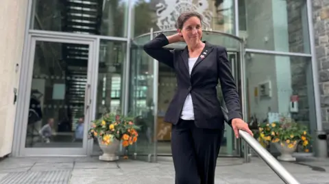 BBC Deputy Lindsay de Sausmarez stands outside a building with a large glass revolving door and several windows. She is holding onto a metal bar at the top of a flight of steps.