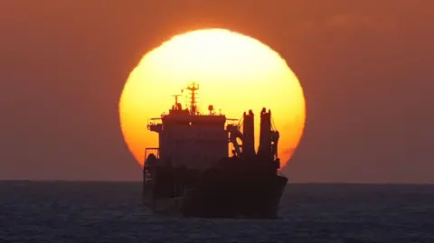 Owen Humphreys/PA Media The sun rises behind a ship in the North Sea off the coast of Whitley Bay. The multi-storey ship is cast in silhouette, but a mast and some work equipment can be made out. The sea is calm and the sky illuminated orange by the sun.