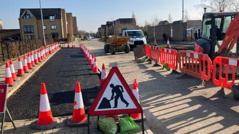 A patch of fresh asphalt surrounded by traffic cones. It is next to an existing road. There is a "Men at Work" road sign at the front of the picture. To the right, on the existing road, there are orange barriers and work vehicles. There are new houses in the distance.