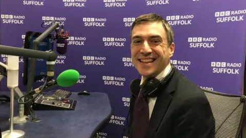 Vikki Irwin/BBC MP Adrian Ramsay is sitting in a radio studio smiling at the camera. He is wearing a suit and dark tie.