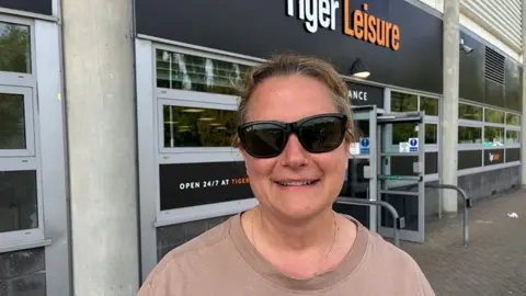 Linsey Smith / BBC Naomi Robinson stands outside the club shop. She has short dark hair and is wearing sunglasses and a beige top.