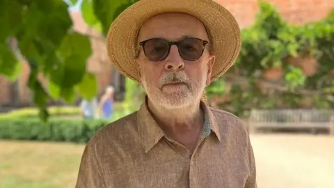 BBC Philip Roberts, a man wearing sunglasses as well as a peach-coloured shirt and a straw hat, is stood in front of an English Tudor building. There is a lawn behind him and a series of low hedges.