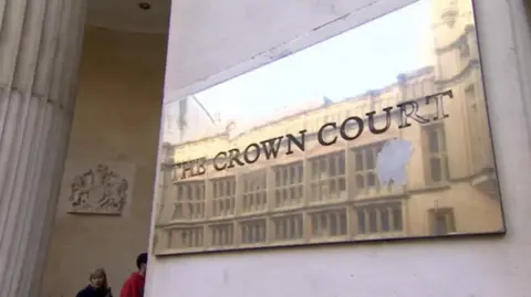 A brass plaque on a sandstone building reading "Crown Court". A column and a court crest is visible in the background. Two people can be seen walking into the building.