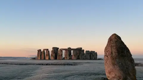 Stonehenge A picture of Stonehenge taken from afar. The top of the sky is blue and fades into an orange at the horizon. The grass surrounding the monument is covered in frost.