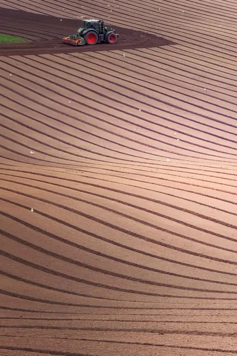 Hamish Spencer-Nairn An aerial view of a tractor with large red wheels ploughing curved rows across a large field, creating sweeping patterns in the soil.