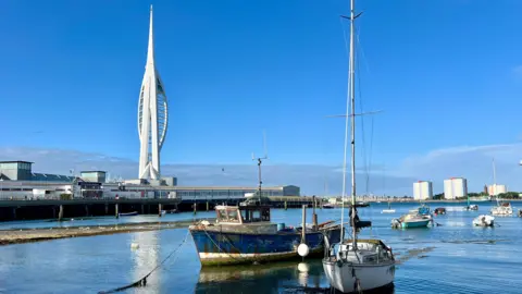 THURSDAY - A view over Portsmouth Harbour as the Spinnaker Tower gleams in the sun over the train station. There are two boats in the foreground, a white sailing boat and a blue fishing boat. Behind several other boats are moored up the harbour. On the far side of the harbour you can see three tower blocks in Gosport 