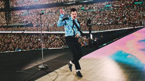 Harry Styles dancing on stage at Wembley wearing a blue floral shirt, black tie, black trousers, white socks and black patent shoes. Thousands of people can be seen standing or watching him from the stands