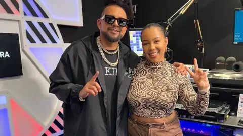 BBC Radio 1Xtra presenter Kaylee Golding in her radio studio stood next to Sean Paul. Kaylee is smiling and has her black hair pulled back. She is wearing a black and white swirly patterned long sleeved top which is slightly cropped and has light brown trousers on. She is doing the peace symbol with her left hand. Sean Paul is stood to her left with his left arm around her shoulder. He has his right arm out and his index finger extended. He has a black T-shirt and black anorak style jacket on and has dark black sunglasses on.