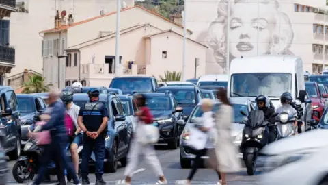 A police officer directs traffic as people walk by and cars wait on a road. A building in the background has artwork depicting Marilyn Monroe