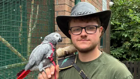 A picure of Connor Atherton who is wearing a hat and green t-shirt and black glasses. He smiles into the camera, whilst holding a gray parrot. 