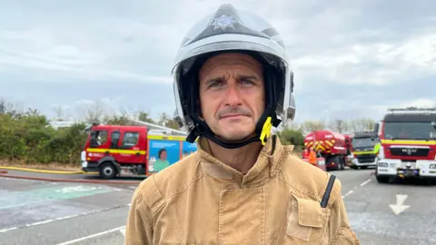 A male firefighter pictured in Weston-super-Mare at the scene of a blaze. He is wearing his uniform, which is brown and includes a helmet. Several fire engines can be seen on the road behind him.