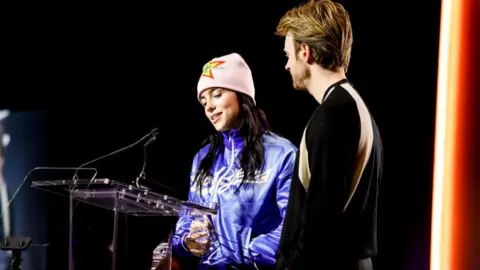 Getty Images Billie Eilish and FINNEAS speak onstage at Variety Hitmakers after being awarded Film Song of The Year/ Billie, a 21-year-old woman, wears a pink beanie hat with a green, yellow and red star on the front. She wears thick rimmed clear glasses and a blue zip-front jacket. Finneas, who looks towards his sister and away from the camera, has strawberry blonde hair and a short beard. he wears a black jumper with camel coloured stripes near the sleeves.