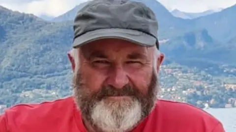A man with a beard smiling at the camera. He is wearing a red t-shirt and has a grey cap on. 