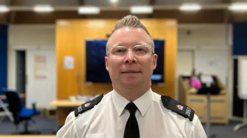 A a policeman with short hair styled so it is spiked up. he is wearing glasses that have translucent frames. He is wearing a white shirt and black tie. A classroom is out of focus behind him. 