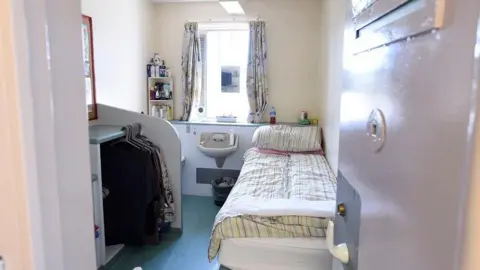 Michael Cooper Open cell in Hydebank. There is a single bed with a duvet on it, a sink, window with curtains and storage area with clothes on hangers. 