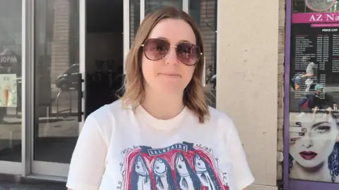 Fiona Callow/BBC A smiling woman looking straight ahead. She has shoulder length, brown hair and is wearing sunglasses and a white T-shirt with fish drawings on the front.
