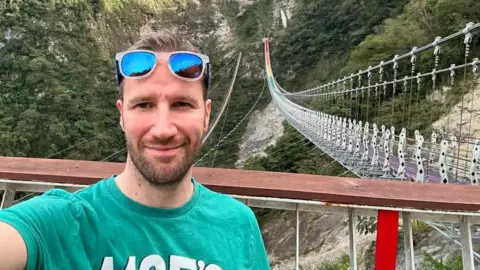 Kevin Smith has sunglasses resting on his forehead and a short beard, and short dark hair. He is standing by a railing, with a long suspended foot bridge to the right, which runs across a large ravine, with a cliff covered in trees beyond.