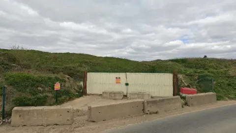 Google Street View Roadside image of the site entrance, with concrete blocks and metal fencing.