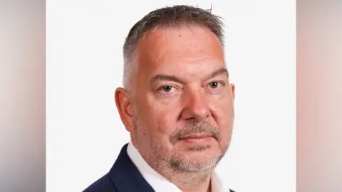 Simon has grey hair and grey and brown facial hair. He is wearing a white shirt and a dark suit jacket. It is a head shot and he is standing in front of a white background