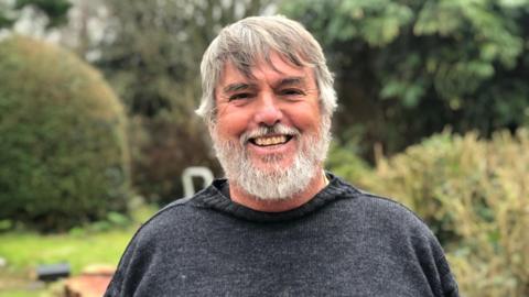 A man with a grey beard wearing a Guernsey-style jumper stands smiling in a garden.
