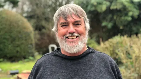 BBC A man with a grey beard wearing a Guernsey-style jumper stands smiling in a garden.