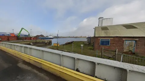 Bentinck Dock at King's Lynn in Norfolk. Shows a grey and yellow metal bridge, a brick building to the right, which is next to a body of water. There is a large container ship in the distance. 