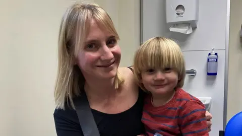 BBC Laura and Sasho in a doctor's evaluation room. A sink with paper towels and soap can be seen behind them. Laura is blonde and wearing a black top. Her arm is around Sasho. Sasho is also blonde sat on Laura's knee, wearing a red and blue stripe jumper. Both of them are looking directly at the camera and smiling.