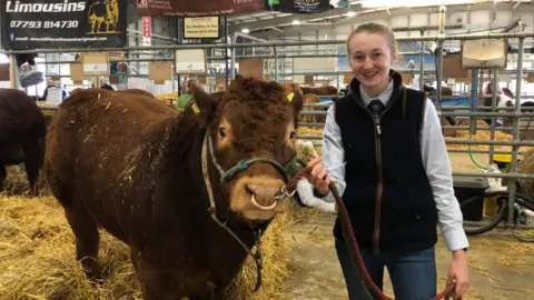 BBC A young woman standing next to a cow at the Balmoral Show, inside a large hanger-like indoor area