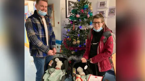 Family photo Ruth and Gavin Coleman standing in front of a Christmas tree smiling, each holding one twin in a baby carrier