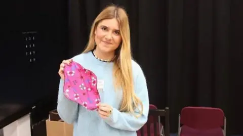 Ella Lambert Ella Lambert holding a reusable period pad which is pink with flowers on it. She has blonde hair and is wearing a blue jumper and smiling at the camera