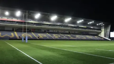A side of a stadium reading "Warriors" across the seats, with a grass pitch below.