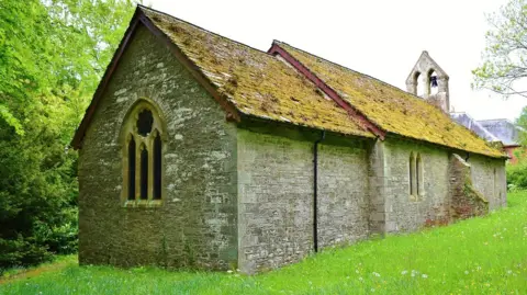 The stone church stands in an area of grass. It has a mossy roof and a window at one end.